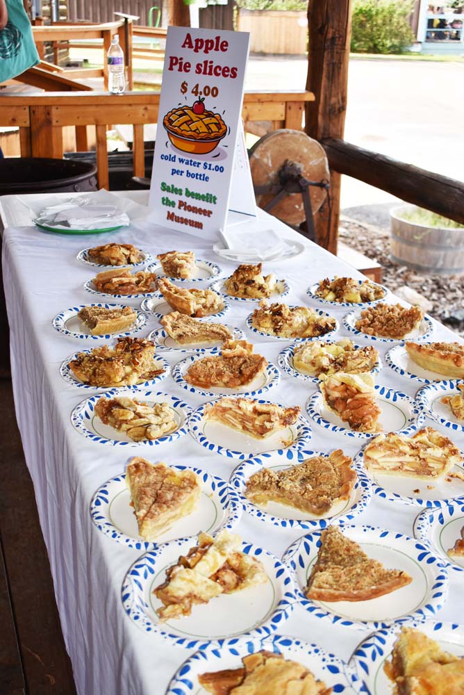 Plates filled with slices of apple pie arranged on a long table covered with a white tablecloth, with a colorful sign reading “Apple Pie slices $4.00” that notes sales benefit the Pioneer Museum. The scene is set on a wooden porch with rustic beams and a glimpse of the museum grounds in the background.