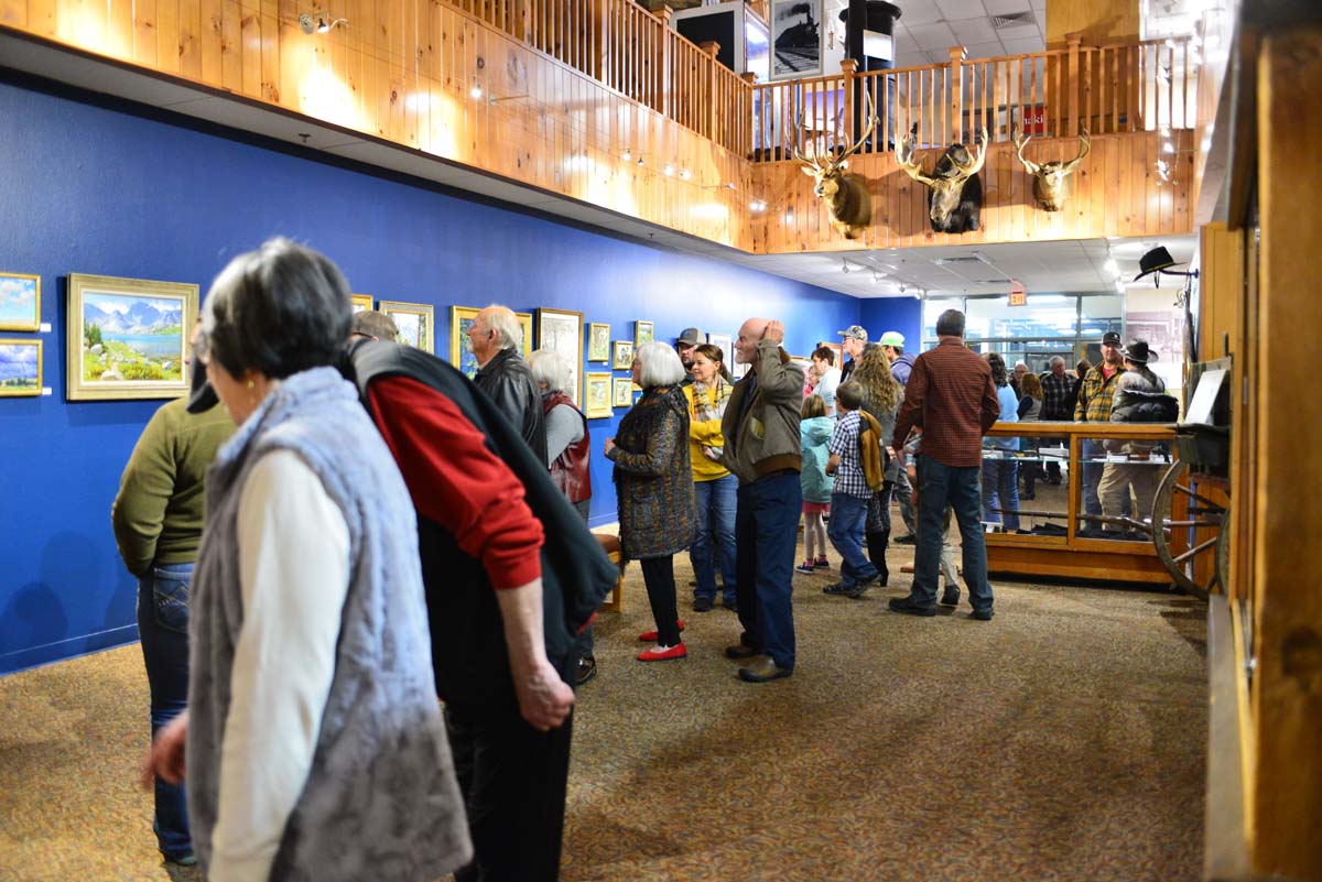 Visitors of all ages fill a brightly lit art gallery at one of the Wind River museums, viewing landscape paintings along a vivid blue wall beneath a warm wood balcony lined with mounted wildlife and historic displays.